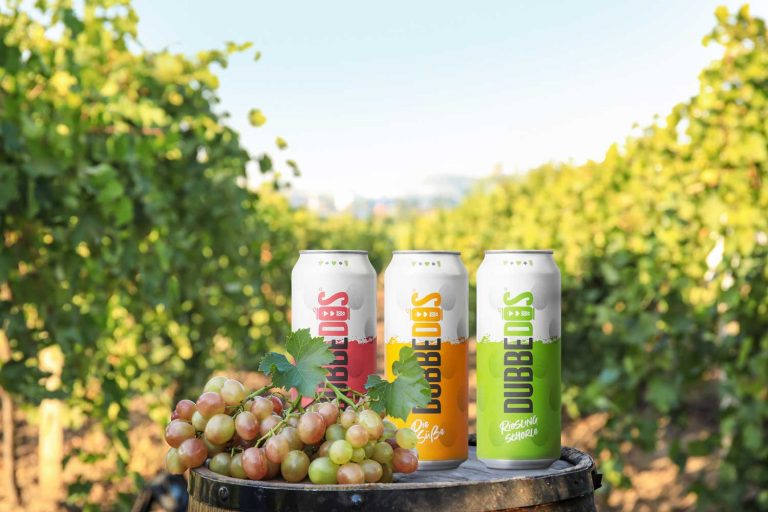 Drei DUBBEDOS Weinschorle-Dosen in verschiedenen Geschmacksrichtungen – Rot, Gelb, und Grün – auf einem Fass, umgeben von frischen Weintrauben, vor dem Hintergrund eines sonnigen Weinbergs.