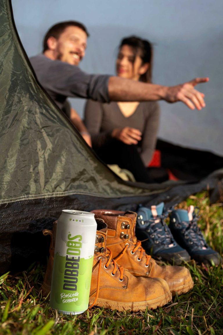 Eine DUBBEDOS-Dose neben einem Paar Wanderschuhe vor einem Zelt auf einer Wiese. Im Hintergrund sind zwei Personen im Zelt zu sehen, die eine entspannte Festivalstimmung genießen.