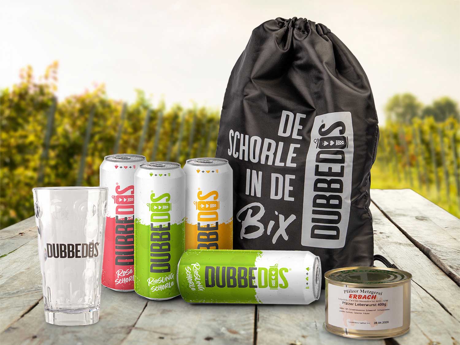 DUBBEDOS Schorle-Paket mit verschiedenen Sorten Weinschorle in der Dose, einem DUBBEDOS-Glas, einer stylischen "De Schorle in de Bix" Tasche und Pfälzer Leberwurst, präsentiert auf einem Holztisch vor einer Weinberg-Kulisse.